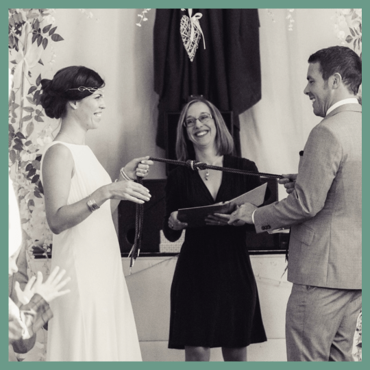 bride and groom smiling as they tie the knot in their handfasting cords in front of their smiling celebrant