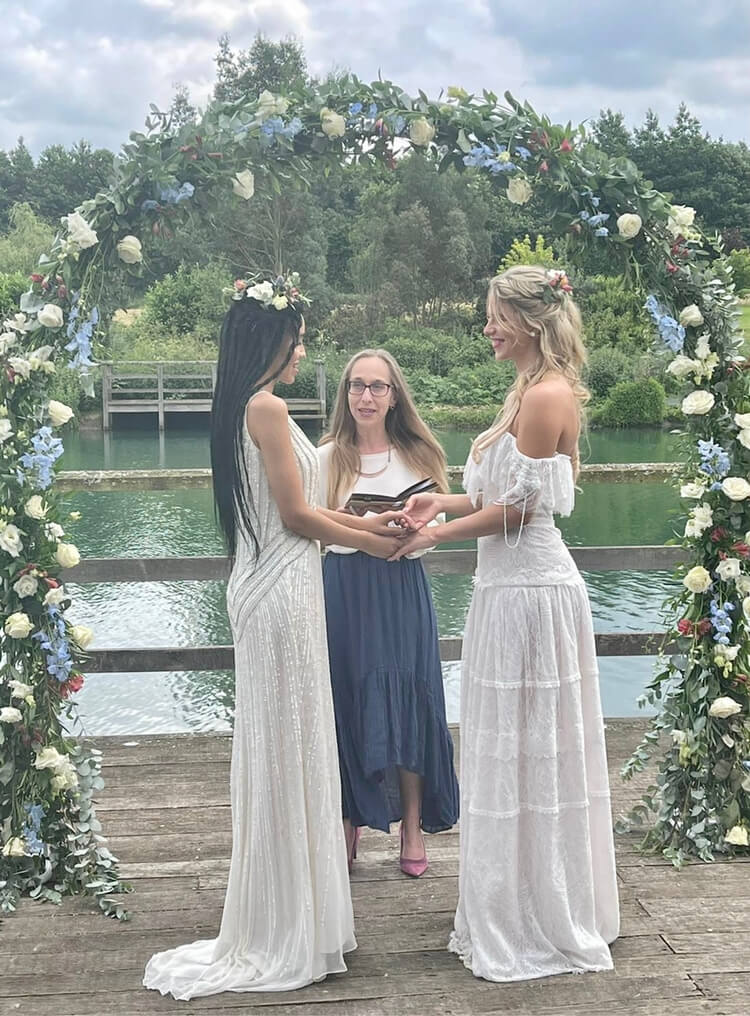 Two brides, one with long black braids and one with lone blonde soft waves, holding hands in front of Alex Hilder - Celebrant under a floral arch by the lake at Kentfields Estate, West Malling, Kent