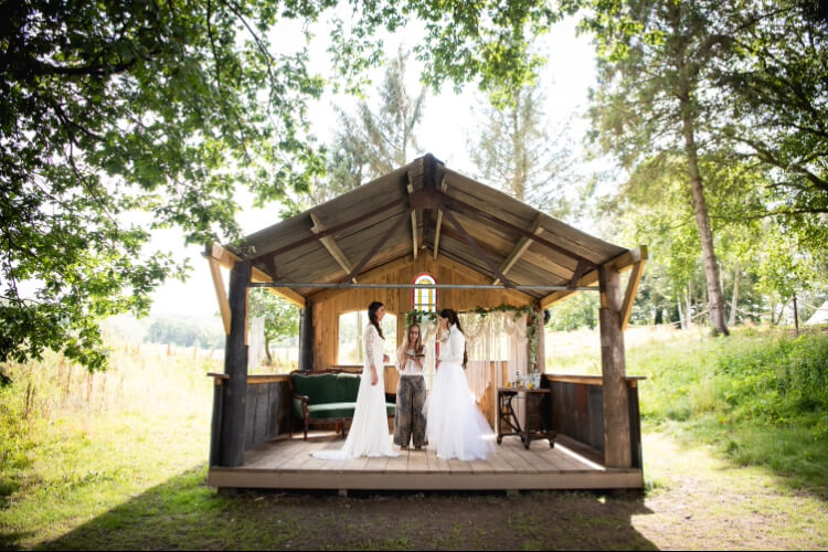 Alex Hilder - Celebrant conducting a ceremony with two brides in the Love Shack at Oak Tree Barn, Tonbridge, Kent, with the sun streaming in from behind them and woodland on either side