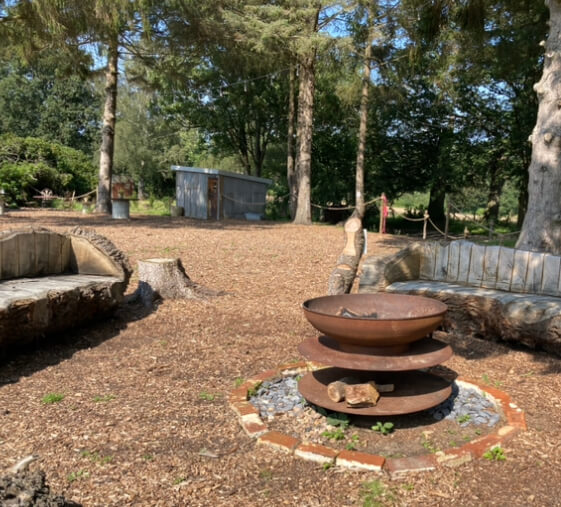 large rusted metal fire bowl stands in a woodland clearing flanked by benches carved from tree trunks at Oak Tree Barn, Tonbridge, Kent