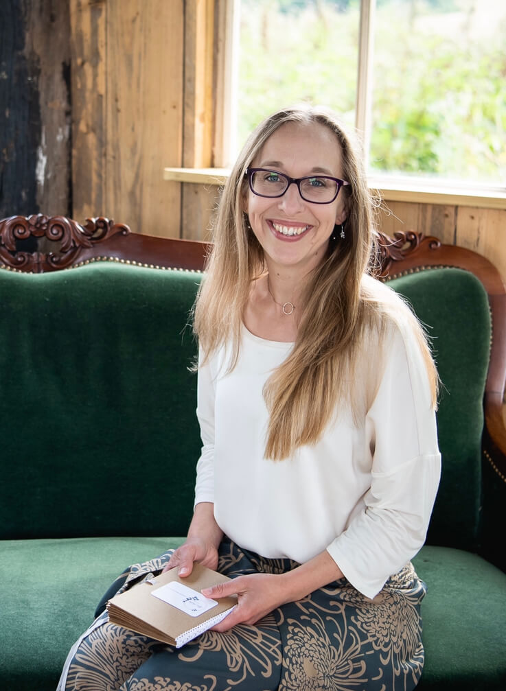 Alex Hilder - Celebrant smiling on a green sofa holding her ceremony book in the outdoor wedding ceremony space at Oak Tree Barn, Tonbridge, Kent