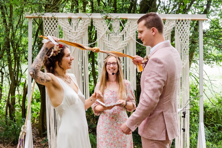 A groom in a pink suit and a bride in a simple white dress pull their handfasting ribbons tight in front of Alex Hilder - Celebrant, who is smiling broadly